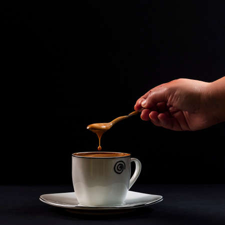 Drop of coffee with milk falling from a spoon held by a person's hand.Photo taken on a black background with artificial light in a studio.It aims to show the concept of creaminess in coffee.の写真素材