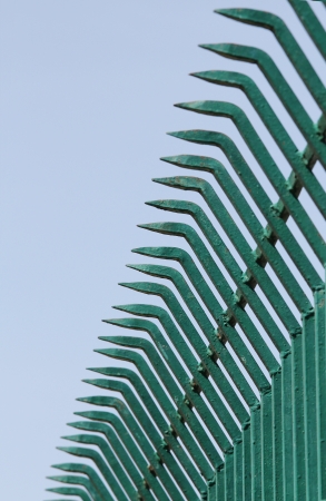 Rows of pointed sharp wrought iron bars forged to form a fence in green color and sky in the backgroundの写真素材
