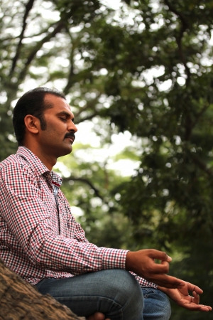 
 a handsome indian businessman meditating under a tree in a garden. The person is sitting in lotus posture and is composed, relaxed closing his eyes for better concentrationの写真素材
