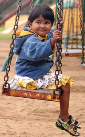 Cute young happy indian girl(kid) playing on a swing in a park. The photo shows summer time playground with a female child smiling and sitting on a swingの写真素材