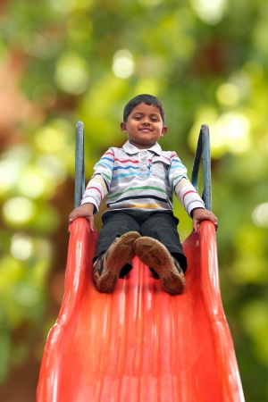 Pretty smiling indian boy(kid) on slider at a park. This boy's photo with green background & clipping path shows summer time playground with a schoolboy playing on a slider の写真素材