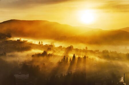 Fantastic sunrise in a mountain village with sunrays passing through a foggy hillsの写真素材