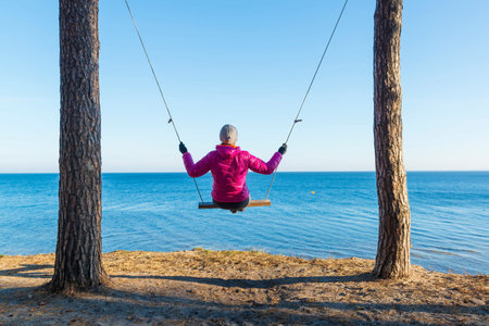 Lonely woman in warm clothes on the swing at the seaside in autumn.