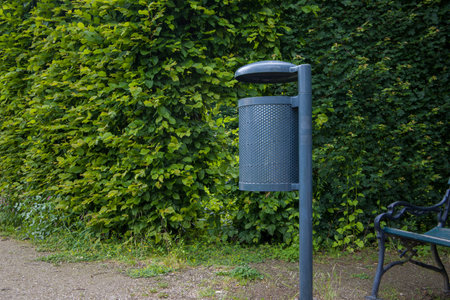 Metal trash can on a park bench in front of a hedge.の写真素材