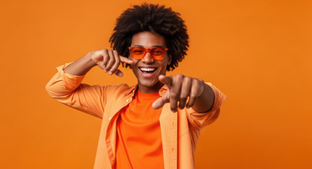 happy african american man in orange sunglasses pointing with fingers at camera isolated on orangeの素材