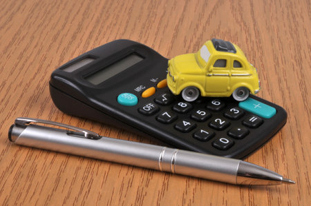 Concept of estimating the price of a vehicle with a small car on a calculator next to a pen on a wooden backgroundのeditorial素材