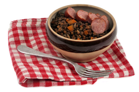 Dish of lentils with slices of smoked sausage served in a bowl with a fork on a checkered napkin on a white backgroundの写真素材
