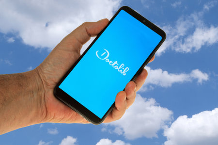 Doctolib application on a smartphone held in hand close-up on a background with a sky and cloudsの写真素材