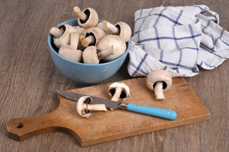 Cut raw button mushroom on a cutting board with a bowl of raw mushrooms and a tea towel in the backgroundの写真素材