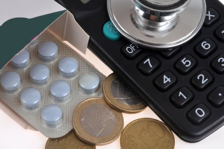 Blister pack of tablets coming out of a box with a stethoscope, calculator and euro coins viewed from above in closeupの写真素材