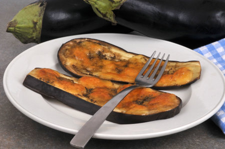 Slices of cooked eggplant with melted cheese on a plate with a fork next to whole raw eggplants in closeupの写真素材