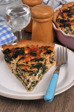 Slice of spinach and salmon savory tart on a plate with a fork and a glass of water closeupの写真素材