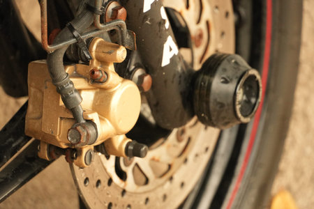 Detailed view of a motorcycle's front wheel brake system. It features a golden caliper and rusty...