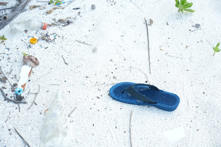 A blue flip flop lies abandoned on a sandy beach littered with plastic waste, including bottles,...