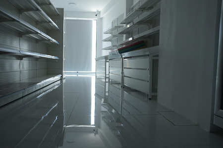 A modern, empty supermarket with white walls, shiny floor, and bare shelves, illuminated by natural...
