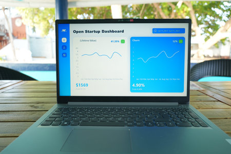 A laptop shows an Open Startup Dashboard with two graphs and key statistics on a wooden table outdoors, with a blurred background of trees and a pool.の写真素材