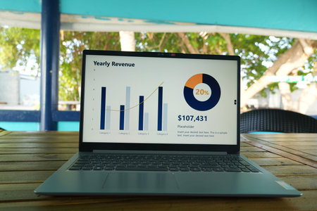A laptop on a wooden table displays a yearly revenue graph and chart, showing business data analysis...