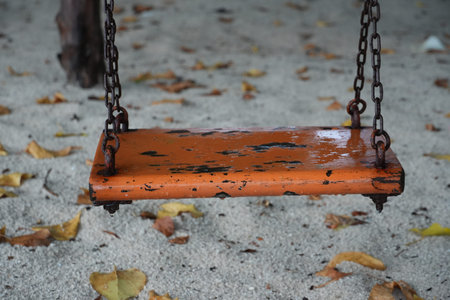An old wooden swing with rusty chains hangs low over sandy ground and dry leaves.