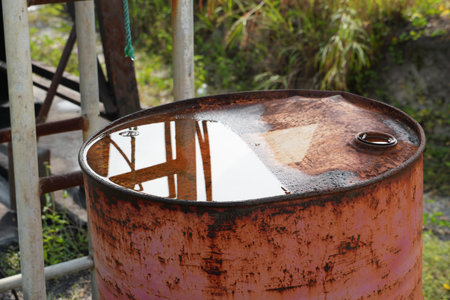A rusty metal barrel filled with rainwater reflects the sky and surrounding industrial elements.の写真素材