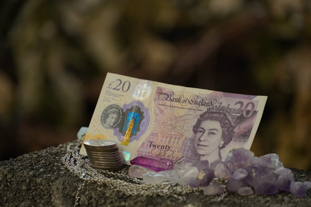 A British twenty-pound note rests with a stack of coins and amethyst crystals on a rough stone.