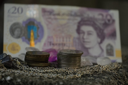 A stack of British coins rests on a silver chain with a blurred twenty pound note behind.の写真素材
