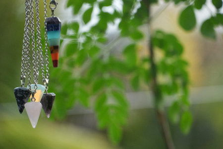 Colorful crystal pendulums with silver chains hang amidst vibrant green leaves.