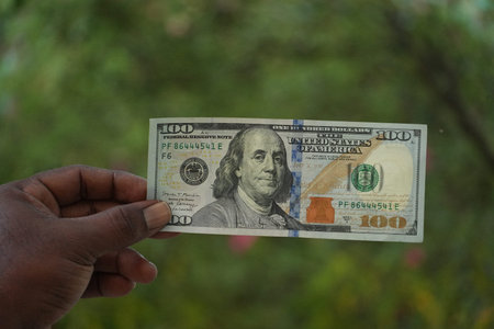 A dark-skinned hand firmly holds a one hundred dollar bill against a blurred green background.
