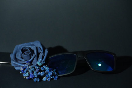 A rich blue fabric rose with small dried flowers rests beside stylish dark sunglasses on a muted background.の写真素材