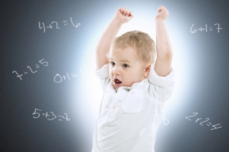 Cute excited little boy standing with his arms raised and mouth open against a school blackboard with simple arithmetic on itの写真素材