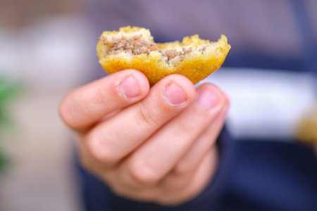 A close-up of a hand holding a partially bitten meat-filled pastry, showcasing the golden crust and...