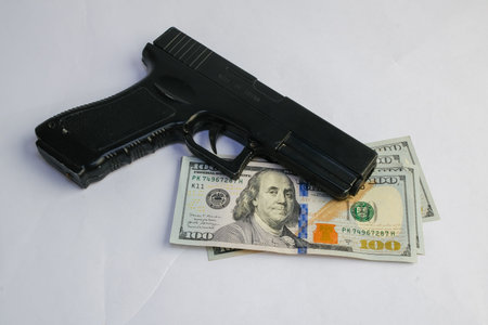 Basra, Iraq - August 5, 2025: A Black Handgun Resting on a Stack of American One Hundred Dollar...