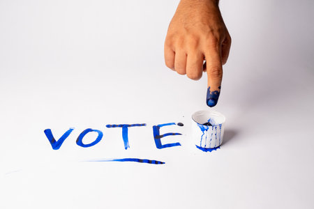 A hand with a blue-painted finger pointing at the word 'VOTE' written in blue paint on a white surface, with a small cup of blue paint nearbyの写真素材