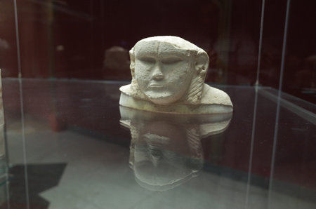 A small stone sculpture of a human face displayed in a glass case, reflecting on the surface belowのeditorial素材