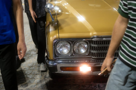 A vintage yellow car with two people standing nearby, one holding a cigarette, at night