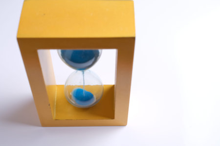 A yellow hourglass with blue sand flowing from the top chamber to the bottom, set against a white...