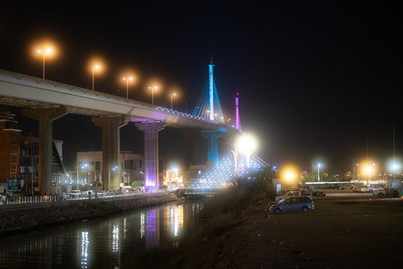 A brightly lit bridge at night with colorful lights, reflecting in the water below, surrounded by buildings and parked carsのeditorial素材