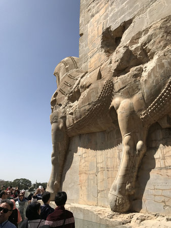 Ancient stone relief of a colossal winged bull with human head (lamassu) carved into a cliff or wall, tourists gathered at base, clear blue sky, sunlit beige limestoneの写真素材