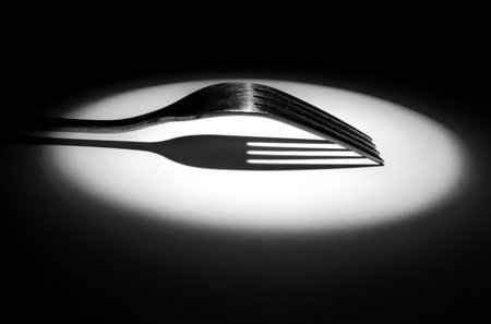 Two metal forks overlapped with dramatic spotlight, forming a curved silhouette and sharp shadow on white surface, black background, high-contrast monochrome still life.の写真素材