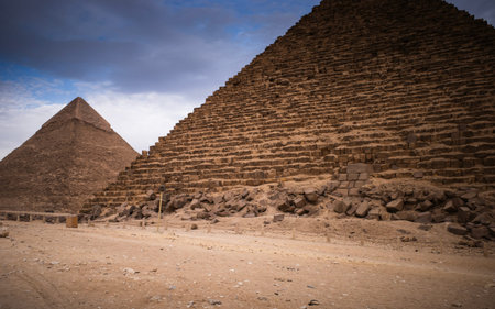 Close-up view of two ancient egyptian pyramids at giza under a moody sky, sandy foreground and scattered stonesのeditorial素材