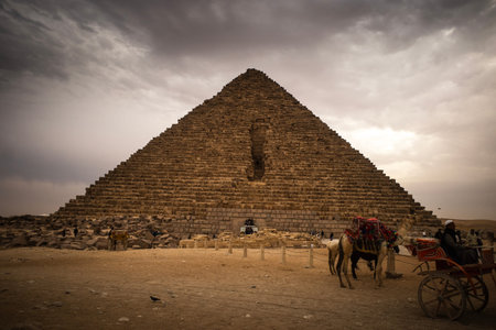 Ancient stone pyramid under moody cloudy sky with camels and a carriage in foreground, desert foreground and scattered rocks, warm tones.のeditorial素材