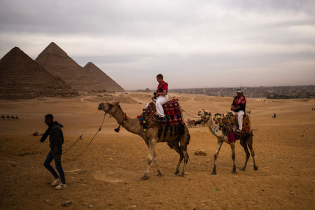 Two camels with riders and a guide cross the sandy Giza plateau with pyramids in the background under an overcast sky.のeditorial素材
