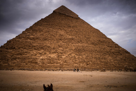 Front view of a large ancient stone pyramid under cloudy sky with sandy foreground and tiny people...