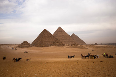 The great pyramids of giza in a vast sandy desert under a cloudy sky, with tourists, camels and...
