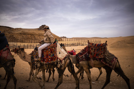 Tourists riding camels with colorful saddles across sandy desert near rock formations under a cloudy...