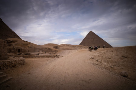 Desert path leading to a lone pyramid under moody cloudy sky, small group of camels and riders near the base, ancient stone structures at left, warm earthy tones.のeditorial素材