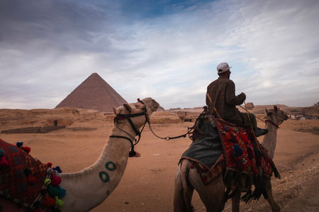 Two camels with a rider in desert near a pyramid under cloudy sky, warm tones, evocative travel...