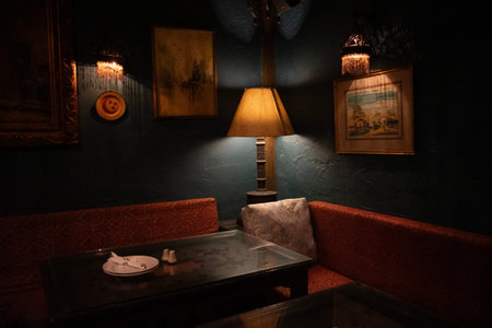Cozy dimly lit corner booth in a vintage bar or cafe with a lamp casting warm light over a table, cushioned red seating, framed artwork on textured dark walls, plate with utensils on tableのeditorial素材