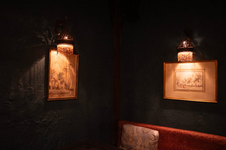 Dimly lit cozy corner with framed sketches on dark textured walls, warm wall sconces casting soft amber light over a cushioned benchのeditorial素材