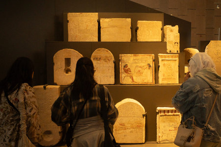Museum visitors viewing ancient stone tablets and reliefs displayed on dark wall shelves in a...