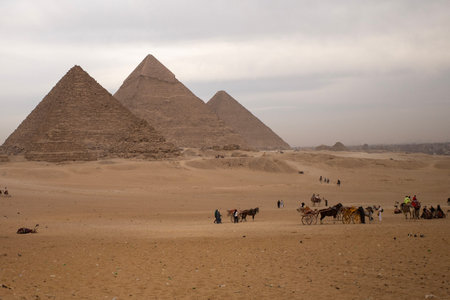 Wide desert scene with the pyramids of giza under overcast sky, small tourists and horse-drawn...